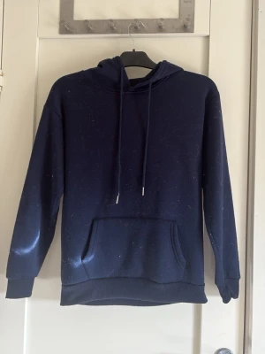 Mörkblå hoodie med ficka - En enkel och stilren mörkblå hoodie med huva och dragsnören. Hoodien har en stor magficka framtill och ribbade muddar vid ärmar och nederkant. Perfekt för en avslappnad och casual look.