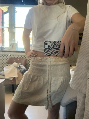 Beige kortkjol med snörning från Zara - Supersöt beige kortkjol med smock i midjan och snörning med pärldetaljer framtill. Kjolen har en lätt volangkant och är gjord i ett mjukt, strukturerat bomullstyg. Perfekt för varma dagar och enkel att styla med en enkel t-shirt.