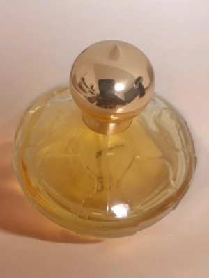 Chopard Casmir - Elegant parfymflaska i glas med rund, platt form och ett glänsande guldfärgat klotformat lock. Flaskan är fylld med gul vätska och har ett dekorativt mönster på glaset. Perfekt som snygg detalj på sminkbordet.