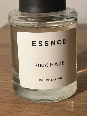 Essnce Pink Haze Eau de Parfum - Minimalistisk parfymflaska från Essnce med doften Pink Haze. Flaskan är cylindrisk i genomskinligt glas med svart lock och vit etikett med svart text. Eau de Parfum-varianten ger en elegant och modern känsla, perfekt för dig som gillar stilrena detaljer. Ny förutom att jag testat den en gång. Jag använder aldrig parfym, detta var en present🙏