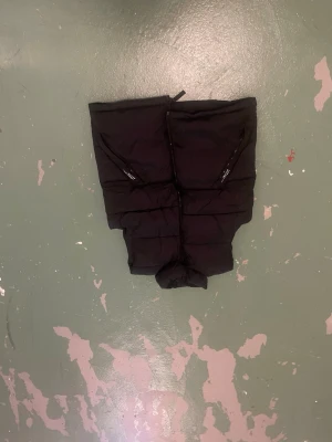 Svart dunväst med dragkedjor - Snygg svart dunväst med två snedställda dragkedjefickor framtill och quiltad design. Västen har hög krage och är vadderad för extra värme. Perfekt att slänga över en hoodie eller tröja för en cool lager-på-lager-look.