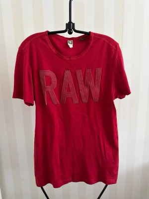 Röd RAW t-shirt från G-Star - Röd t-shirt från G-Star RAW med stora bokstäver 'RAW' framtill. Klassisk rund halsringning och korta ärmar. Tillverkad i mjuk bomull som känns skön mot huden. Perfekt för en avslappnad och trendig look.