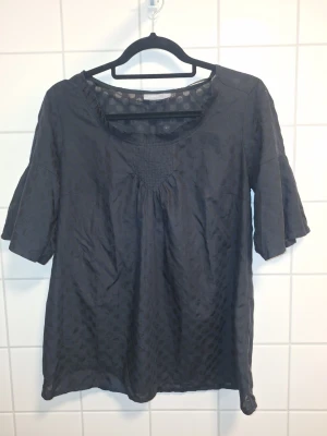 Tunn svart prickig blus med trekvartsärm - Snygg svarr blus med diskret prickigt mönster och trekvartsärmar. Blusen har halsringning med smockdetalj framtill och ett lätt tunt tyg med vävda prickar som ger en cool look. Perfekt att styla med jeans eller kjol.