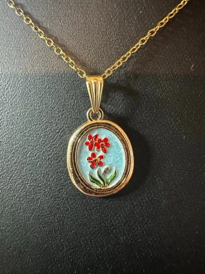 Guldigt halsband med blommigt hänge - Unikt halsband med tunn guldfärgad kedja och ovalt hänge. Hänget har en ljusblå bakgrund med röda blommor och gröna blad i emalj. Perfekt för dig som gillar färgglada detaljer och vintagekänsla. Kedjan är tunn och smidig, och hänget ger en personlig touch till din stil.