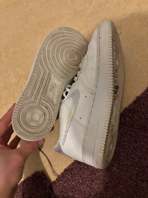 Nike Air Force 1 med holografisk swoosh - Säljer ett par vita Nike Air Force 1 sneakers med holografisk swoosh och detaljer på hälen. Klassisk siluett med perforerad tå och tjock sula. Användt dom bara några gånger nästa helt nya 