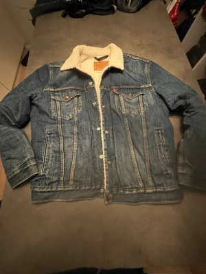 Levi's fodrad jeansjacka blå M - Klassisk blå jeansjacka från Levi's med vit teddyfoder och snygga detaljer. Jackan har två bröstfickor med knappar, sidofickor och metallknappar framtill. Perfekt oversized look och ikonisk Levi's-lapp i nacken.