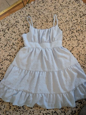 Ljusblå kort klänning med volanger xs - Supersöt ljusblå klänning med tunna axelband och volangkjol. Klänningen har ett diskret prickigt mönster i tyget och markerad midja. Perfekt för varma dagar och ger en fräsch och somrig look.