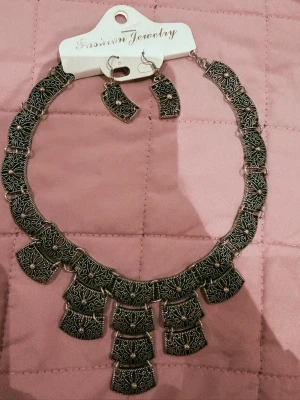 Antiksilverfärgat statementhalsband - Snyggt halsband i antiksilverfärg med matchande örhängen. Halsbandet består av flera rektangulära länkar med detaljerade mönster och små pärlliknande dekorationer. Perfekt för att lyfta en outfit med en bohemisk touch.