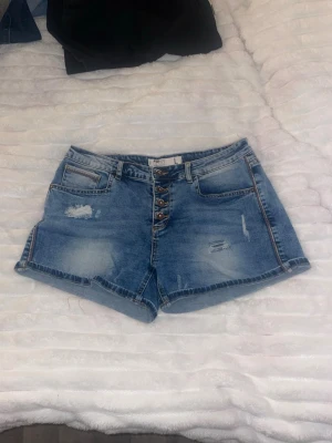 Blå denimshorts från FB Sister - Säljer ett par blå denimshorts från FB Sister med slitna detaljer och knappar framtill. Shortsen har klassisk femficksdesign och är tillverkade i bomull. Perfekta för sommaren och ger en avslappnad look.