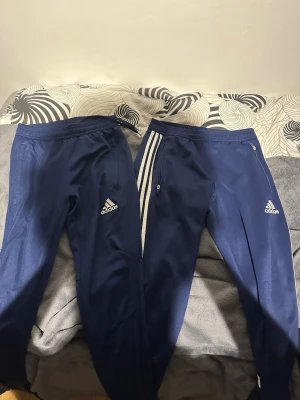Mörkblå Adidas mjukisbyxor - Två par mörkblå mjukisbyxor från Adidas med klassiska vita ränder längs sidorna och Adidas-logga på låret. Byxorna har resår i midjan och smal passform. Perfekta för chill eller träning.