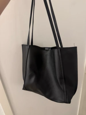 Svart shoppingväska i skinnimitation - En stilren svart shoppingväska med dubbla axelband och enkel design. Väskan är tillverkad i skinnimitation och har en rymlig insida, perfekt för att bära med sig allt man behöver. Passar till många olika outfits och är lätt att matcha.