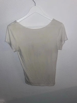 Vit basic t-shirt med rosett på ryggen - En enkel vit t-shirt från lager 157 med rund halsringning och korta ärmar. Tillverkad i mjuk bomull som känns skön mot huden, och som är öppen i ryggen och sen en rosett man kan knyta baktill. Perfekt att matcha med jeans eller shorts för en clean och avslappnad stil.
