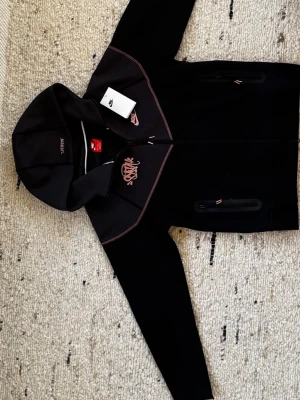 Nike Tech x Syna Wrld - Svart-grå Nike Tech med huva och dragkedja framtill. Det är en collab mellan Nike och Syna Wrld. Jackan har rosa detaljer och logga på bröstet samt två fickor med dragkedja. Prislapp tillkommer vid köp.