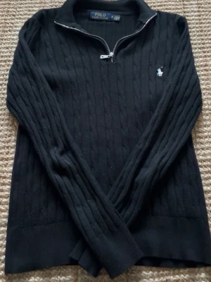 Svart kabelstickad tröja Polo Ralph Lauren - Kabelstickad svart tröja från Polo Ralph Lauren med halv dragkedja och klassisk vit broderad logga på bröstet. Tröjan har ribbade muddar och hög krage, perfekt för lager på lager. Materialet är mjukt och håller värmen.
