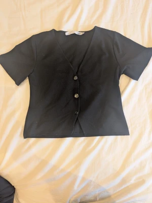 Svart kortärmad topp från 157 - Stilren svart topp från lager 157 med v-ringning och tre knappar framtill. Kortärmad modell i mjukt material som sitter snyggt och passar till det mesta. Enkel och clean design som är lätt att matcha.