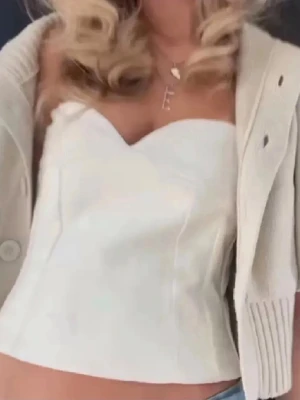 Vit korsettopp med hjärtformad urringning - Snygg vit korsettopp med hjärtformad urringning och figurnära passform. Toppen är axelbandslös och har en croppad längd som passar perfekt till jeans eller kjol. Materialet känns slätt och har en lätt glansig finish.