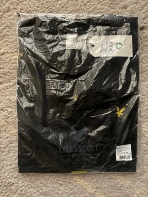 Svart t-shirt från Lyle & Scott - Snygg svart t-shirt från Lyle & Scott med klassisk gul logga på bröstet. Tillverkad i ekologisk bomull för en skön och hållbar känsla. Enkel och stilren design som passar till allt.