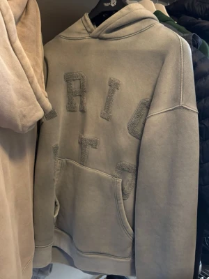 Beige hoodie från Axel Arigato - Snygg beige hoodie från Axel Arigato med stora, broderade bokstäver på bröstet. Hoodien har en klassisk känguruficka och huva. Tillverkad i mjuk bomull för en avslappnad och trendig look. Det är storlek  M. Och den är lite mer baggie i storleken.