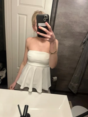 Vit bandeau topp med utsvängd midja - Säljer en snygg vit bandeau topp med utsvängd midja. Toppen är axelbandslös och har en figurnära passform upptill som sedan faller ut i en volangliknande form. Perfekt för dig som vill ha en stilren och trendig look.