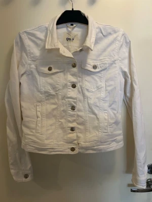 Vit jeansjacka från ONLY - Stilren vit jeansjacka från ONLY med klassisk krage, silverfärgade knappar och två bröstfickor med lock. Figurnära passform och långa ärmar med knapp vid ärmslut. Perfekt att slänga över en t-shirt för en fräsch look.