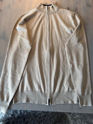 Beige ziptröja med hög krage - Snygg beige ziptröja med hög krage och hel dragkedja framtill. Tröjan är tunnstickad och har långa ärmar samt ribbade muddar vid ärmslut och nederkant. Perfekt för lager på lager och enkel att matcha med olika outfits. Aldrig använd
