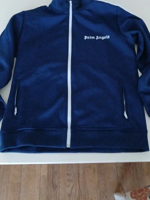 Blå ziptröja från Palm  - Snygg blå ziptröja från Palm med vit dragkedja och logga på bröstet. Tröjan har hög krage och två fickor med dragkedja. Perfekt för en streetwear-look och enkel att matcha med andra plagg.