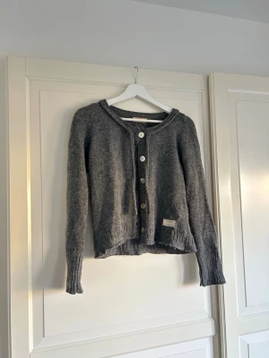 Grå stickad kofta med knappar - Mysig grå stickad kofta med rund hals och långa ärmar. Koftan har ribbade muddar, knäppning framtill med knappar. Använd en del men fortfarande i ett fint skick!!