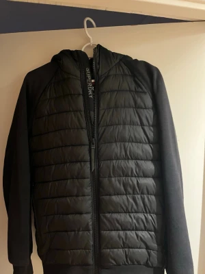 Svart pufferjacka från Superdry - Säljer en svart pufferjacka från Superdry med huva och dragkedja. Jackan har quiltad framsida och rygg, samt släta ärmar och muddar. Märkeslogga syns på både fram- och baksidan. Perfekt för kalla dagar och riktigt snygg streetstil.