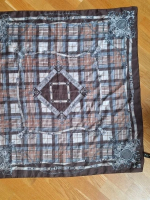 Brunrutig scarf från TieRack, ArtOfScarf - Säljer en snygg brunrutig scarf från TieRack, tillverkad i Italien. Sjalen har klassiskt rutmönster i brunt, beige och grått med dekorativa detaljer i hörnen och längs kanterna. Materialet är 100% polyester och den är mjuk och lätt att styla.