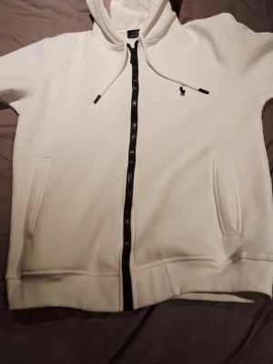 Vit hoodie från Polo Ralph Lauren - Snygg vit hoodie från Polo Ralph Lauren med dragkedja framtill och svart broderad logga på bröstet. Hoodien har två fickor, ribbade muddar och justerbar huva med snören. Perfekt för en clean och stilren look.