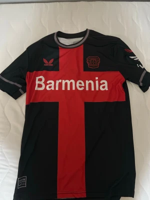 Bayer Leverkusen matchtröja svart/röd - Snygg Bayer Leverkusen matchtröja från Castore i svart med röd korsdesign och vita detaljer. Tröjan har klubbmärke och sponsortryck på bröstet samt rund halsringning med grå kant. Perfekt för fotbollsfans och träning.