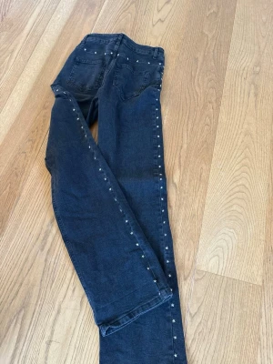 Mörkgrå jeans med stjärnnitar straight leg. - Snygga mörkgrå jeans med raka ben och silverfärgade stjärnnitar längs sidorna och baktill vid linningen. Jeansen har klassisk femficksmodell och bälteshällor. Perfekta för dig som vill ha en unik detalj på dina jeans.