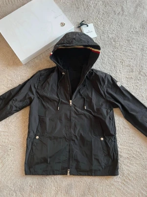 Svart vindjacka från Moncler - Snygg svart vindjacka från Moncler med huva och dragkedja framtill. Jackan har två framfickor med tryckknappar och en ikonisk Moncler-logga på ärmen. Huvan har en randig detalj i rött, vitt och blått. Tillverkad i lätt, vattenavvisande material.Helt ny med etiketter, om du har några frågor tveka inte att fråga. Priset kan diskuteras.