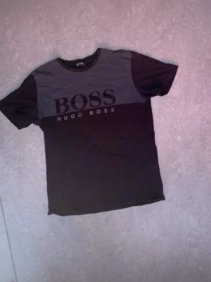 Svart och grå Hugo Boss t-shirt - Snygg svart och grå t-shirt från Hugo Boss med stor BOSS-logga och texten HUGO BOSS på bröstet. Klassisk rund hals och korta ärmar. Perfekt för dig som gillar stilrena märkeskläder med tydlig logga.