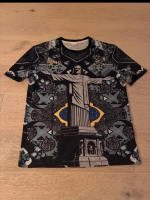 Svart Brasil t-shirt med Kristusstatyn - Unik svart t-shirt med stort tryck av Kristusstatyn i Rio de Janeiro och detaljerade grafiska mönster i grått, blått och gult. Texten 'BRASIL' i guld på bröstet. Perfekt för dig som gillar statement-plagg och coola prints.