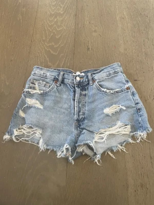 Blå slitna jeansshorts med fransar - Säljer ett par blå denimshorts med hög midja och råa, fransiga kanter. Shortsen har flera slitningar framtill och klassisk femficksdesign. Perfekta för sommaren och ger en avslappnad, trendig look.