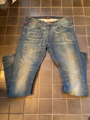 Vintage lee jeans - Säljer ett par blå jeans med snygga slitningar och tvättade detaljer. Klassisk femficksmodell med orange sömmar och knappgylf. Jeansen har en rak passform och är tillverkade i denim. Bra skick och storlek 31/32