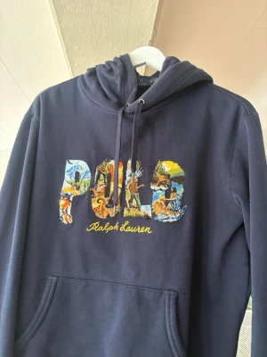 Marinblå hoodie Polo Ralph Lauren - Snygg marinblå hoodie från Polo Ralph Lauren med stor färgglad logga på bröstet och gul brodyrtext. Klassisk känguruficka och justerbar huva med dragsko. Tillverkad i mjukt material som känns skönt mot huden.