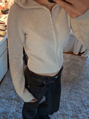 Beige zip hoodie med huva - Mysig stickad beige hoodie med dragkedja framtill och huva. Tillverkad i ett mjukt, fluffigt material som ger en avslappnad och trendig look. Modellen är croppad och har långa ärmar med ribbade muddar. Sitter som en s/xs. Har noppror men det går enkelt att lösa med noppermaskin.