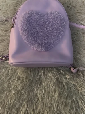 Ljuslila ryggsäck med fluffigt hjärta - Söt ljuslila ryggsäck med ett stort fluffigt hjärta på framsidan. Väskan har släta axelband och dragkedja upptill. Materialet är syntetiskt läder och hjärtat är gjort av mjukt, lurvigt tyg. Perfekt för dig som gillar gulliga detaljer och pastellfärger.