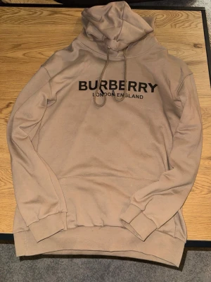 Beige hoodie från Burberry - Snygg beige hoodie från Burberry med svart logotyp och texten 'London England' på bröstet. Hoodien har huva med dragsko och ribbade muddar vid ärmar och nederkant. Tillverkad i mjukt bomullsmaterial som känns skönt mot huden.
