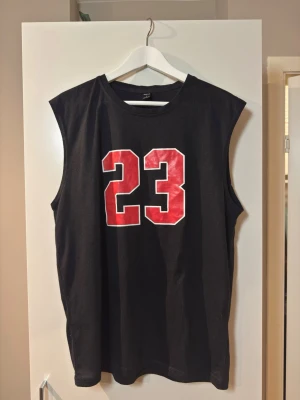Svart ärmlös topp med röd 23-print - Cool svart ärmlös topp med stort rött nummer 23 framtill. Oversized passform och rund halsringning. Perfekt för en sportig och avslappnad stil. Materialet känns mjukt och bekvämt, troligen bomull eller en bomullsblandning.