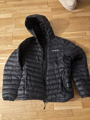 Svart dunjacka med huva PeakPerformance - Snygg svart dunjacka från PeakPerformance med huva och dragkedja framtill. Jackan har två sidofickor med dragkedja och som ger en sportig look. Perfekt för kalla vinterdagar och enkel att matcha med olika outfits.