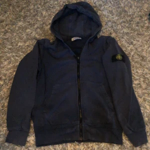 Blå hoodie från Stone Island - Säljer en mörkblå hoodie från Stone Island med klassisk patch på ärmen. Tröjan har huva, dragkedja framtill och två fickor. Tillverkad i mjuk bomull och har långa ärmar med muddar. Perfekt för dig som gillar streetwear och vill ha något stilrent. 