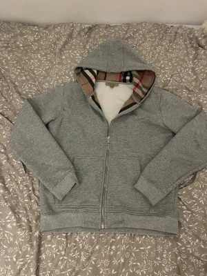 Grå hoodie från Burberry med dragkedja stolek M lite mindre  - Snygg grå hoodie från Burberry med klassiskt rutigt foder i huvan och dragkedja framtill. Hoodien har långa ärmar, två fickor och broderad Burberry-logga på bröstet. Perfekt för en avslappnad och stilren look.