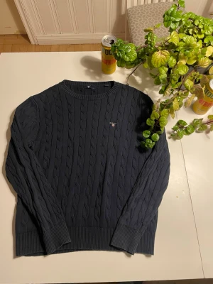 Mörkblå kabelstickad tröja GANT - Säljer en mörkblå kabelstickad tröja från GANT med rund halsringning och broderad logotyp på bröstet. Tröjan har långa ärmar och ribbstickade muddar vid ärmslut och nederkant. Perfekt för dig som gillar klassisk och stilren look.