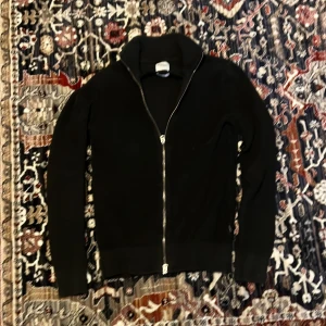 Svart ribbad ziptröja från Dobber - Säljer en svart ribbad ziptröja från Dobber med hög krage och hel dragkedja framtill. Tröjan har långa ärmar och breda muddar vid ärmslut och nederkant. Perfekt för en stilren och enkel look. Går gärna ner i pris