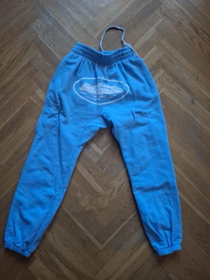 Blå mjukisbyxor med märke Corteiz  - Blå mjukisbyxor med resår i midjan och benslut. Byxorna har ett vitt tryck på framsidan och är tillverkade i mjukt bomullstyg. Perfekta för en avslappnad stil.