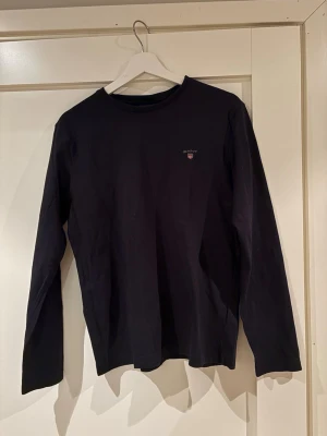 Svart långärmad tröja från Gant - En stilren svart långärmad tröja från Gant med rund halsringning och diskret logga broderad på bröstet. Tillverkad i mjuk bomull som känns skön mot huden. Perfekt att matcha med jeans eller chinos för en clean look.