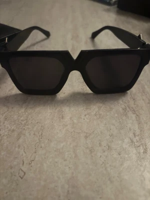 Svarta solglasögon  - Snygga svarta solglasögon  med rektangulära bågar och mörka glas. På sidorna finns en guldfärgad detalj. Perfekt accessoar för dig som vill sticka ut med en trendig look.